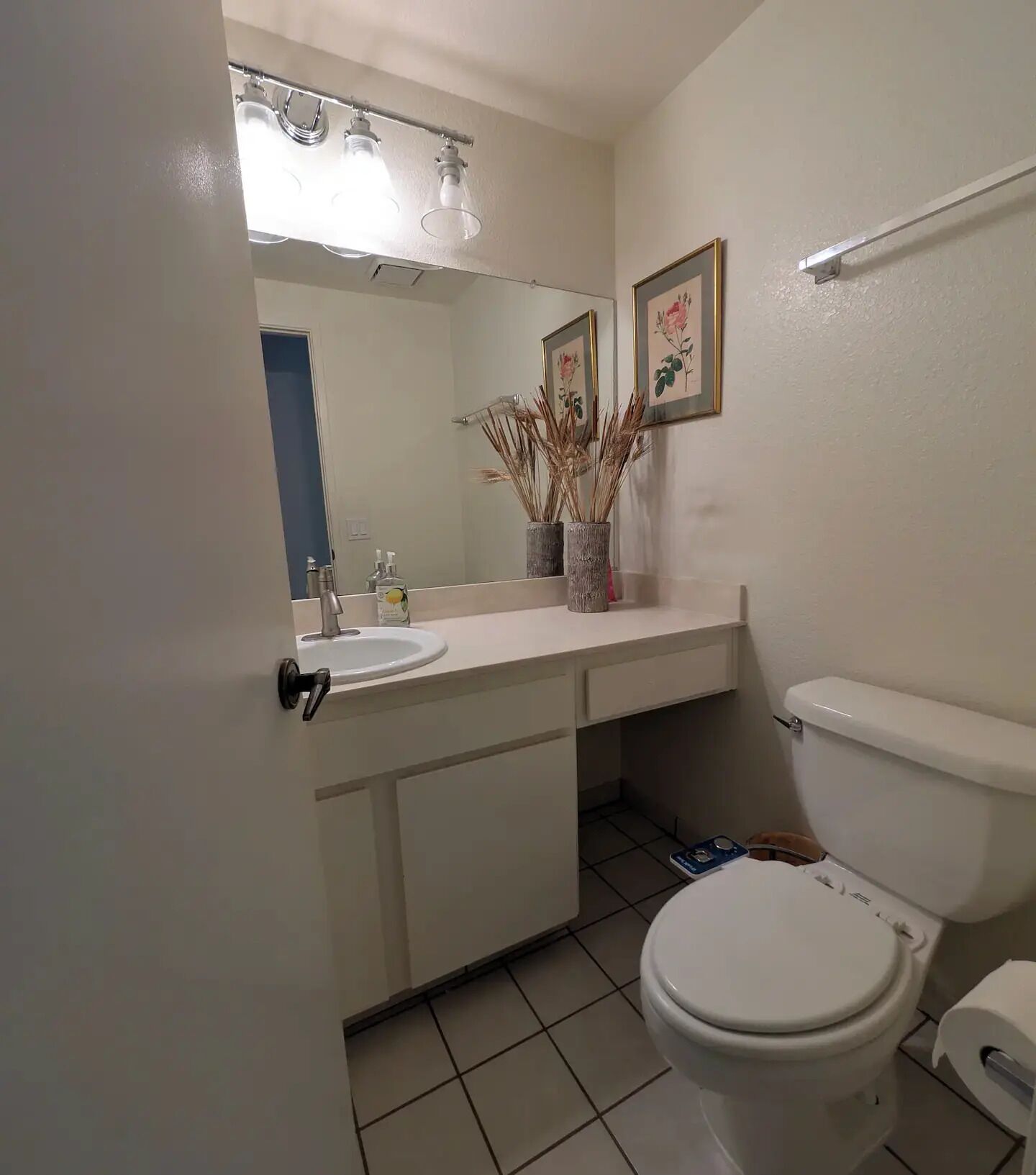 A bathroom with a white sink, mirror, and toilet. There are two framed pictures on the wall, a vase with dried plants, and a towel rack.