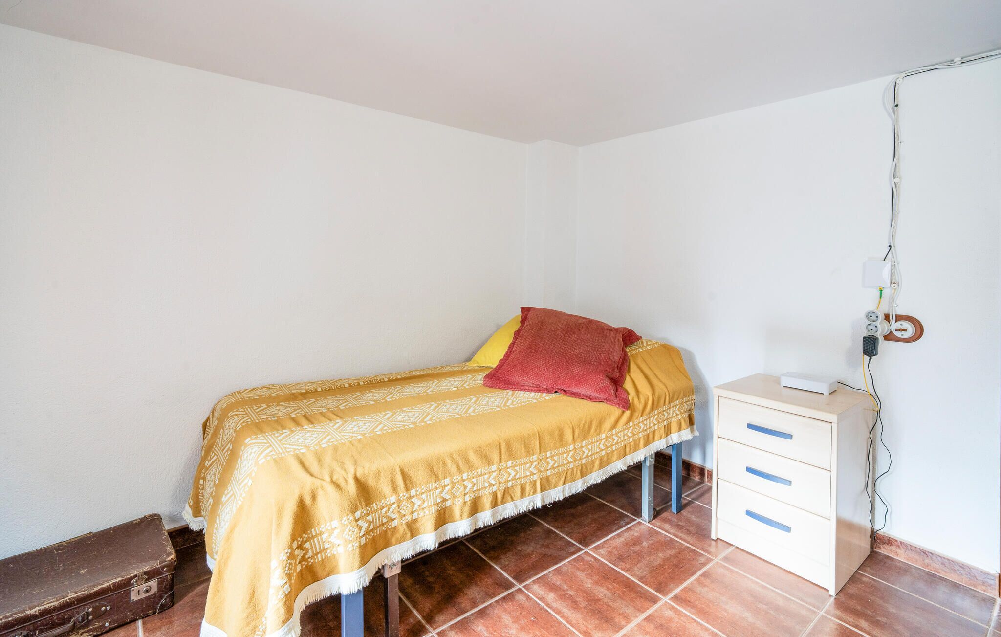 Una habitación pequeña con cama individual, colcha estampada, almohada roja, una maleta y un escritorio con cajones.