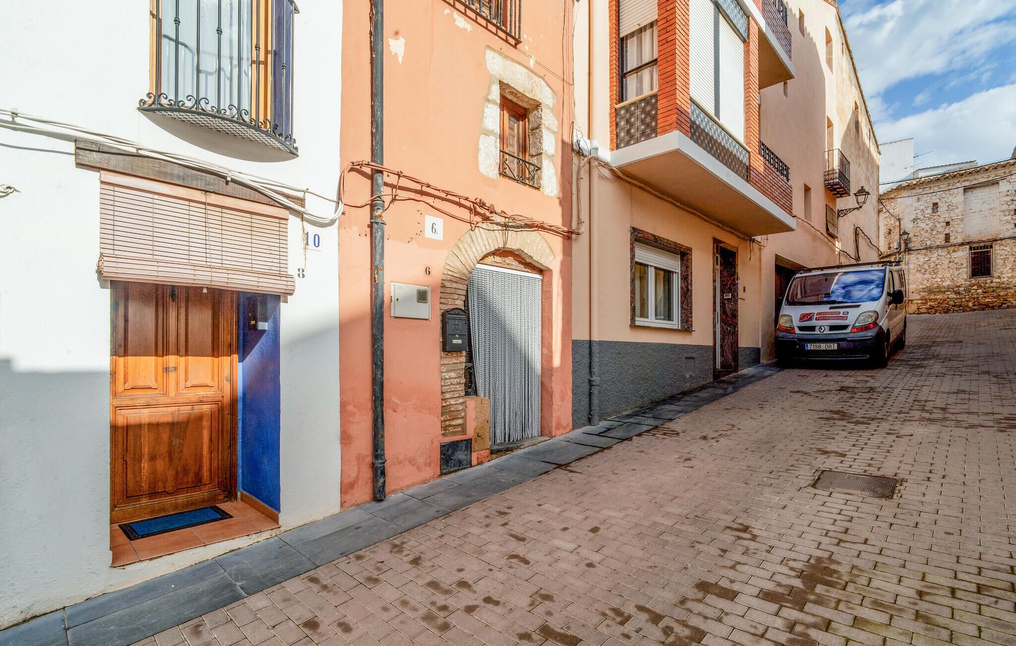 A narrow cobblestone street with buildings on either side. There is a wooden door on the left and a vehicle parked on the right.