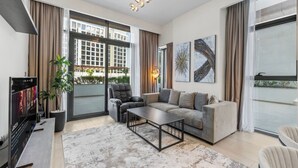 Ein modernes Wohnzimmer mit einer Couch, Couchtisch und Fernseher. Große Fenster mit Vorhängen, eine Pflanze und Kunstwerke an den Wänden.