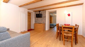 A cozy living room with a sofa, wooden dining table, and chairs. There's a stone fireplace and a TV mounted on the wall. - Appartamento Focobon (Falcade)
