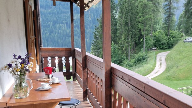 A wooden deck with a table and chairs, a vase of flowers, and a view of a forested mountain area. - Appartamento Focobon (Falcade)