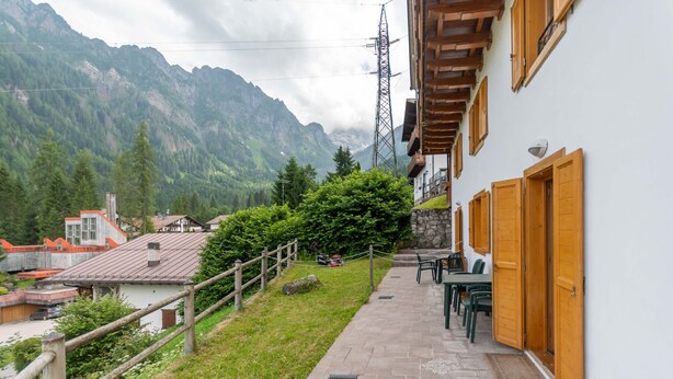 A pathway leading to a building with wooden doors and a stone wall, surrounded by greenery and mountains in the background. - Casa Alessandra 1 (Falcade)