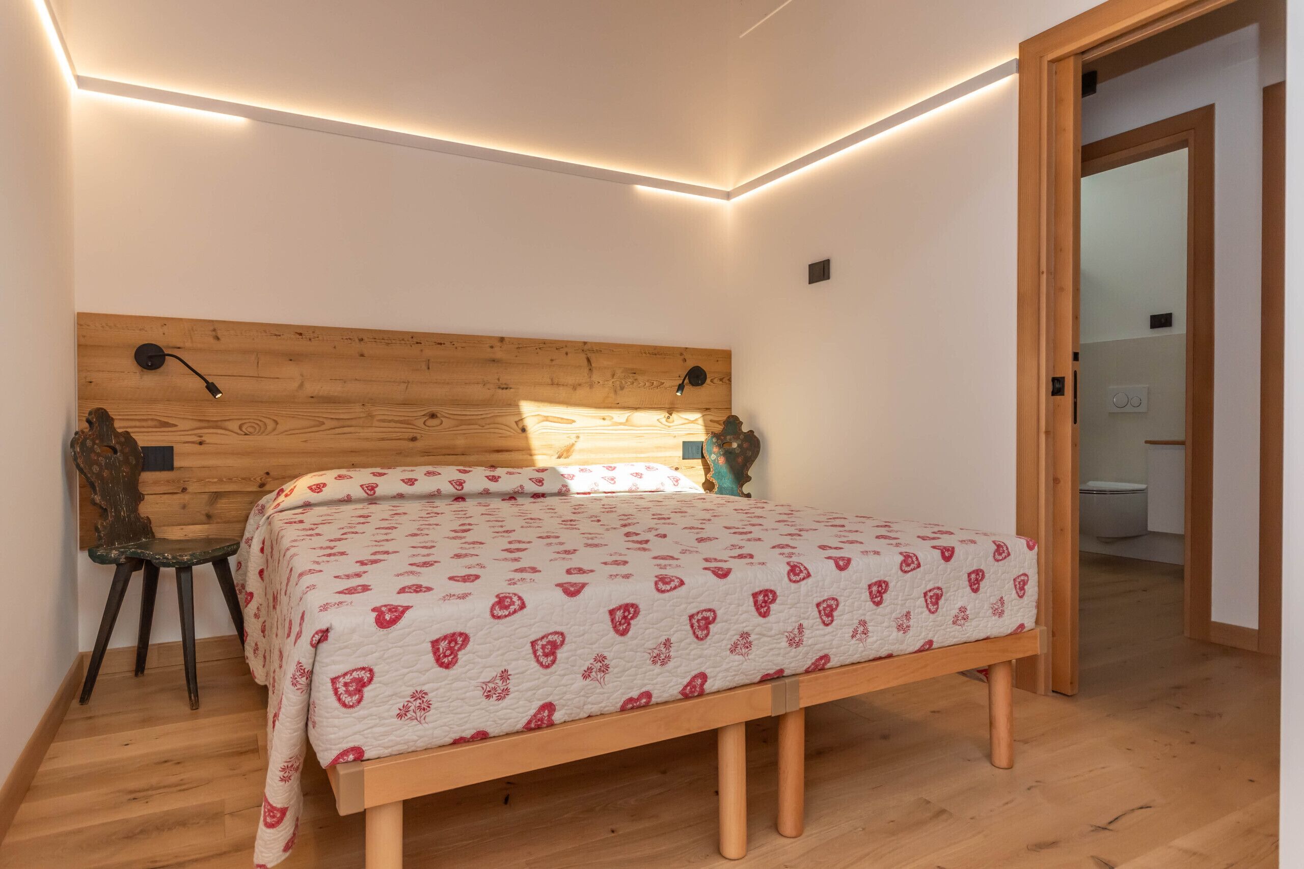 Un dormitorio con cabecera de madera, cama con colcha estampada, mesita de noche, una silla y piso de madera.