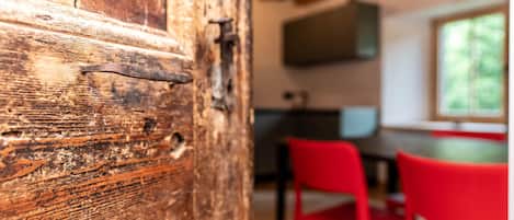 A rustic wooden door with metal hinges, a kitchen area with a TV mounted above a counter, and red chairs in the background.