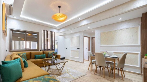 A modern living room with a sectional sofa, a dining area with a table and chairs, and a large window providing natural light. - Cosy new apartment Gueliz (Marrakech)