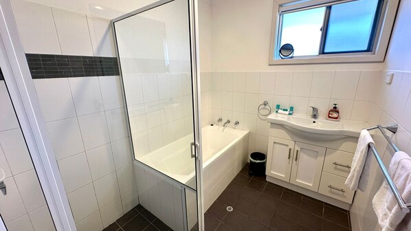 A modern bathroom with a glass shower enclosure, a bathtub, a sink with toiletries, and a window above the sink.