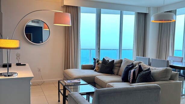 A modern hotel room with a large window, a sofa, a coffee table, and a desk with a lamp.