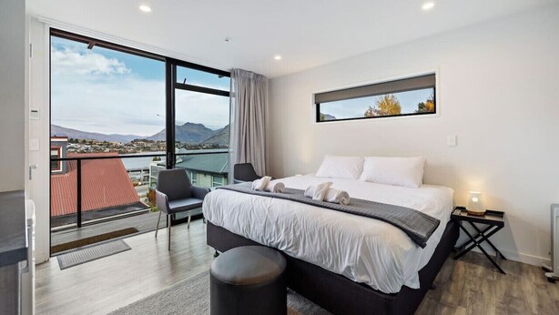 A modern hotel room with a large bed, a chair, a small table with a lamp, and a view of the mountains through large windows. - Frankton Breeze 1 | Close to Queenstown CBD (Queenstown)