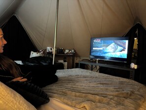 Una habitación con tienda de campaña, una cama, un televisor mostrando 'The Other Woman' y un ordenador portátil sobre una mesa.