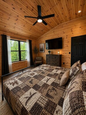 A bedroom with a wooden ceiling, a bed with a quilt, a chair, a TV, and a door. - The Quana -  Hot tub|Huge Entertainment Room (Logan)