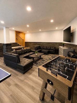 A modern living room with a foosball table, leather sofas, a wooden staircase, and a flat-screen TV.