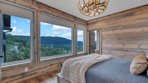 A bedroom with a large window offering a mountain view, wooden walls, and a modern chandelier. - Hang Yer Hat - Breathtaking Views + Elegant Modern Interiors! (Sevierville)