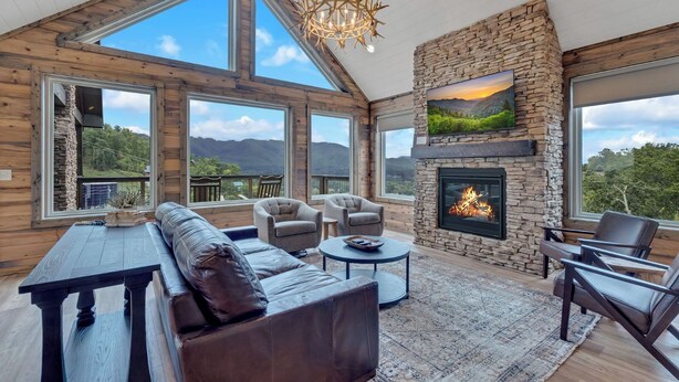 A spacious living room with a stone fireplace, large windows offering mountain views, and a cozy seating area. - Hang Yer Hat - Breathtaking Views + Elegant Modern Interiors! (Sevierville)