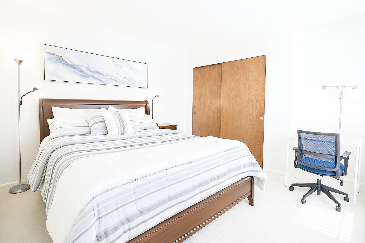 A neatly made bed with a striped bedspread, a wooden wardrobe, a desk with a chair, and a floor lamp.