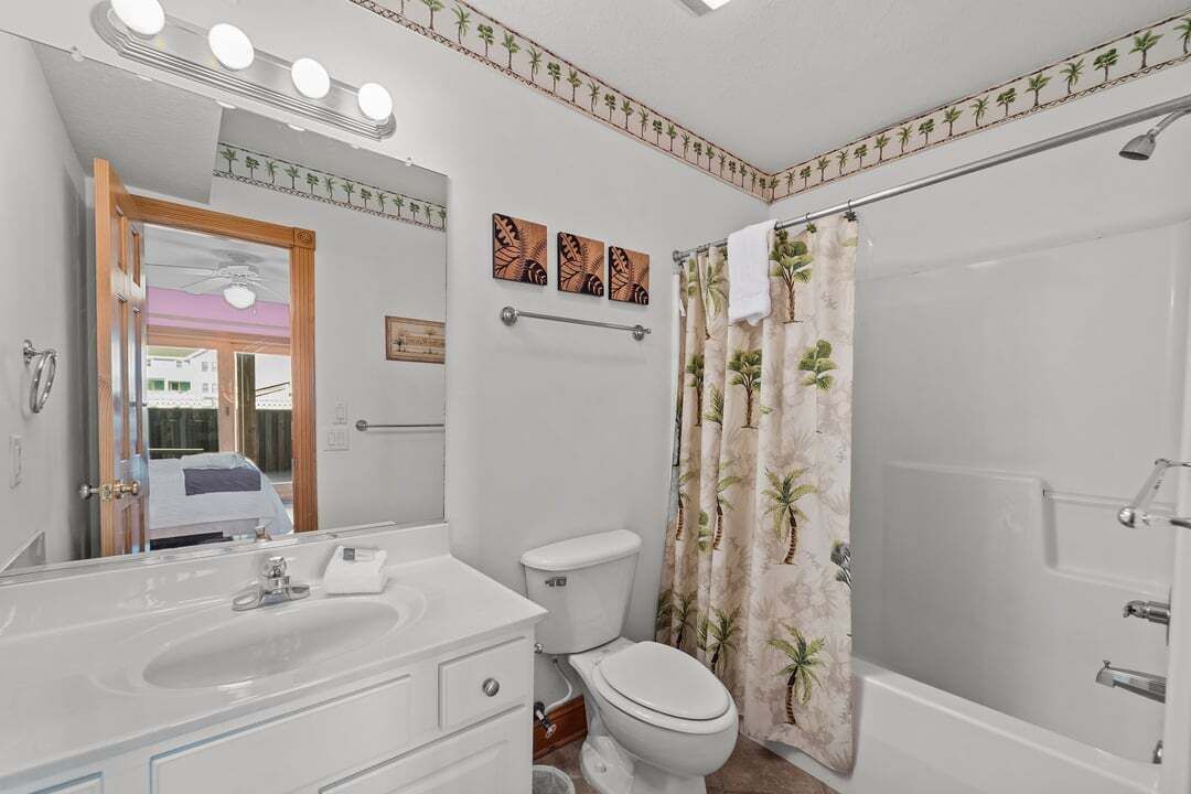 Un baño con lavamanos, inodoro y bañera blancos. Hay tres luces empotradas en la pared sobre el espejo, y un perchero sobre el inodoro.
