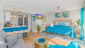 A modern hotel room with a kitchenette, a bed with a blue bedspread, a ceiling fan, and a TV on the wall. - St Clements 704 (Myrtle Beach)