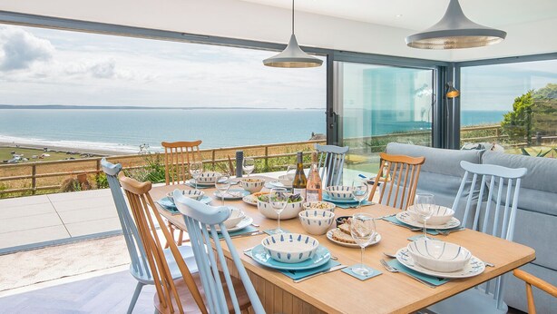 A dining area with a view of the sea, a wooden table set with plates and glasses, and a sofa in the background.