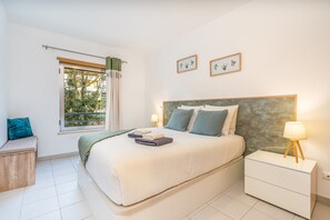 Ein Schlafzimmer mit Bett, Nachttischen, einem Fenster mit Vorhängen und gerahmten Bildern an der Wand.