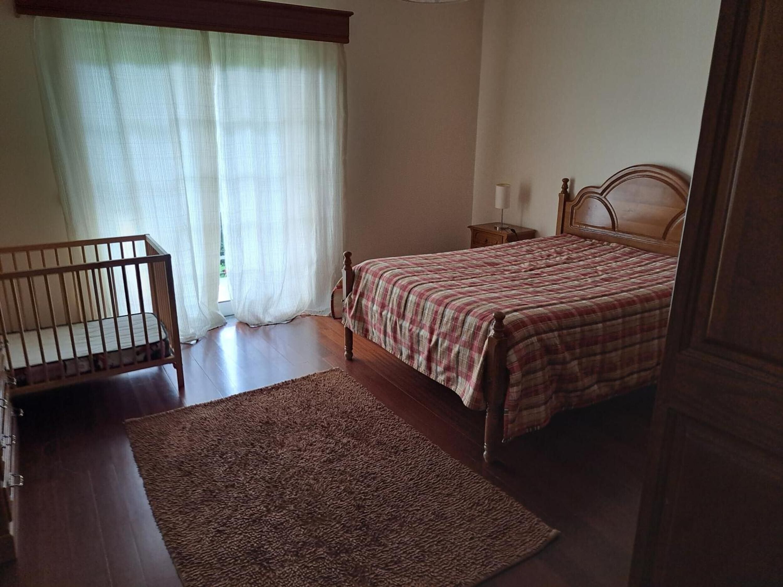 Une chambre à coucher avec un berceau, un lit recouvert d’une couverture à carreaux, une table de chevet et un plancher en bois.