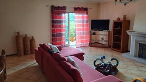 A living room with a sofa, a TV stand, a fireplace, and a view through a window with curtains. - Casa Pinho – Private Jacuzzi, Mountain View & Fireplace (São Pedro do Sul)