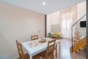 Un coin repas avec une table blanche, des chaises en bois et une assiette de fruits. Un escalier mène à un espace de vie comprenant un canapé et des rideaux.