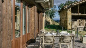 Ein Holzhaus mit großen Fenstern, eine Terrasse mit Tisch und Stühlen und eine Steinstruktur im Hintergrund.