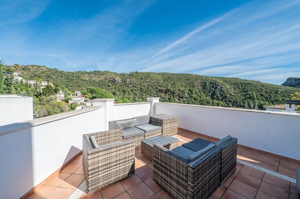 Une terrasse sur le toit, meublée de mobilier en osier, offrant une vue dégagée sur une colline verdoyante et, au loin, un bâtiment blanc.