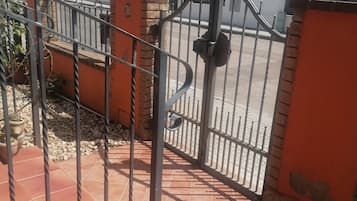 Een hekpoort met een metalen poort, een betegeld terras en een gebouw met een rood dak op de achtergrond.