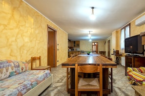 A dining area with a wooden table and chairs, a sofa with a patterned cover, a television, and a kitchen in the background.