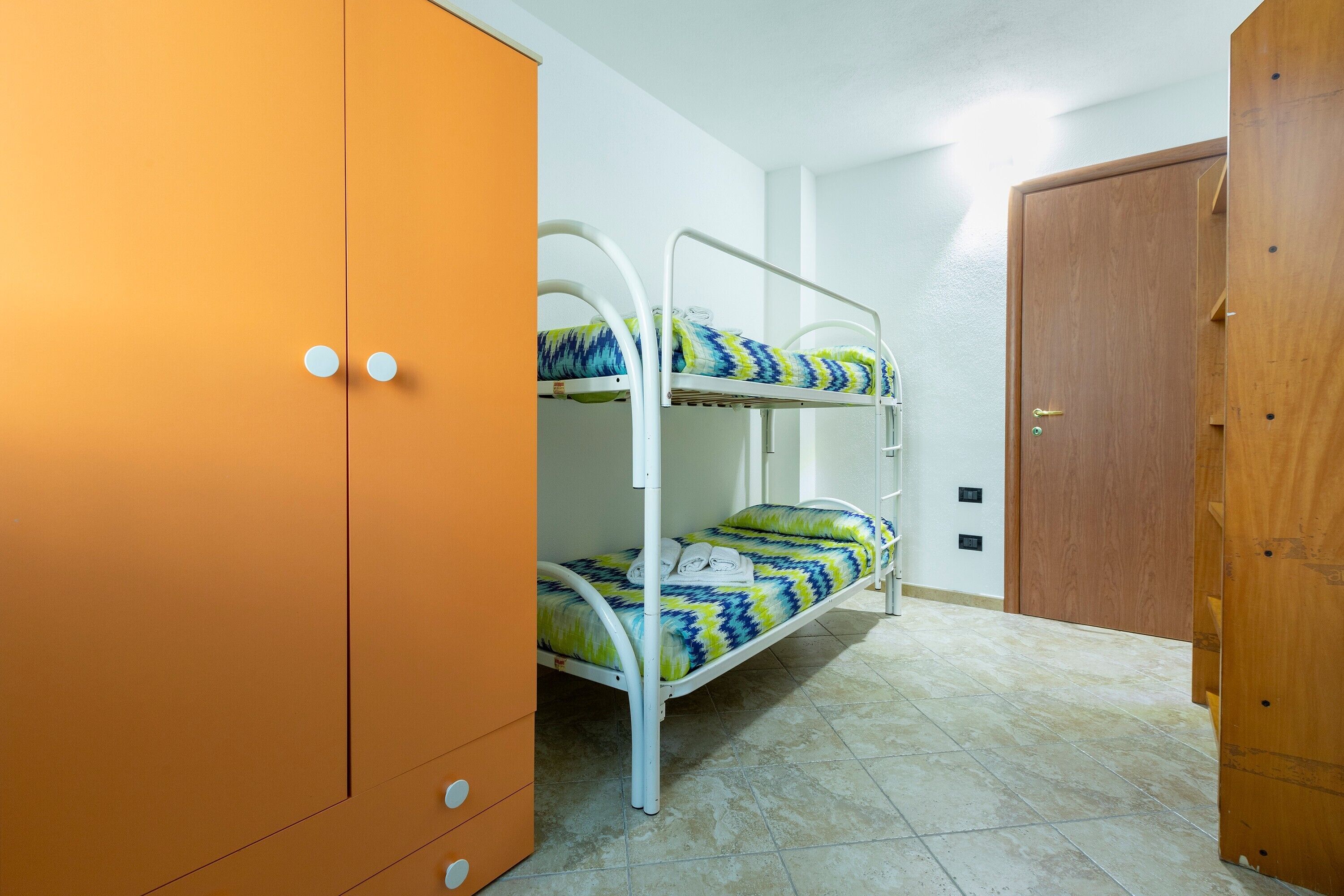 A bunk bed with a patterned bedspread, an orange wardrobe with two drawers, and a wooden door.