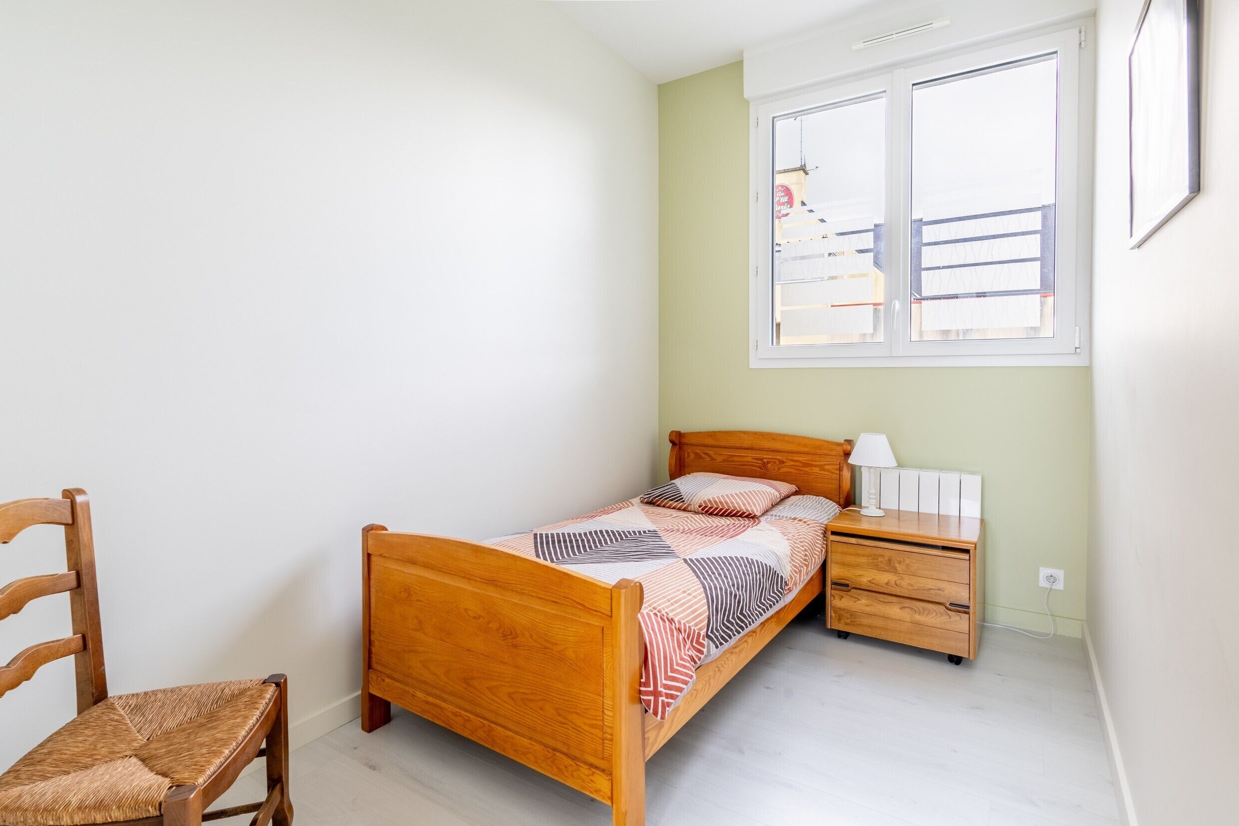 A bedroom with a wooden bed, a nightstand, a chair, and a window with a view of a balcony.