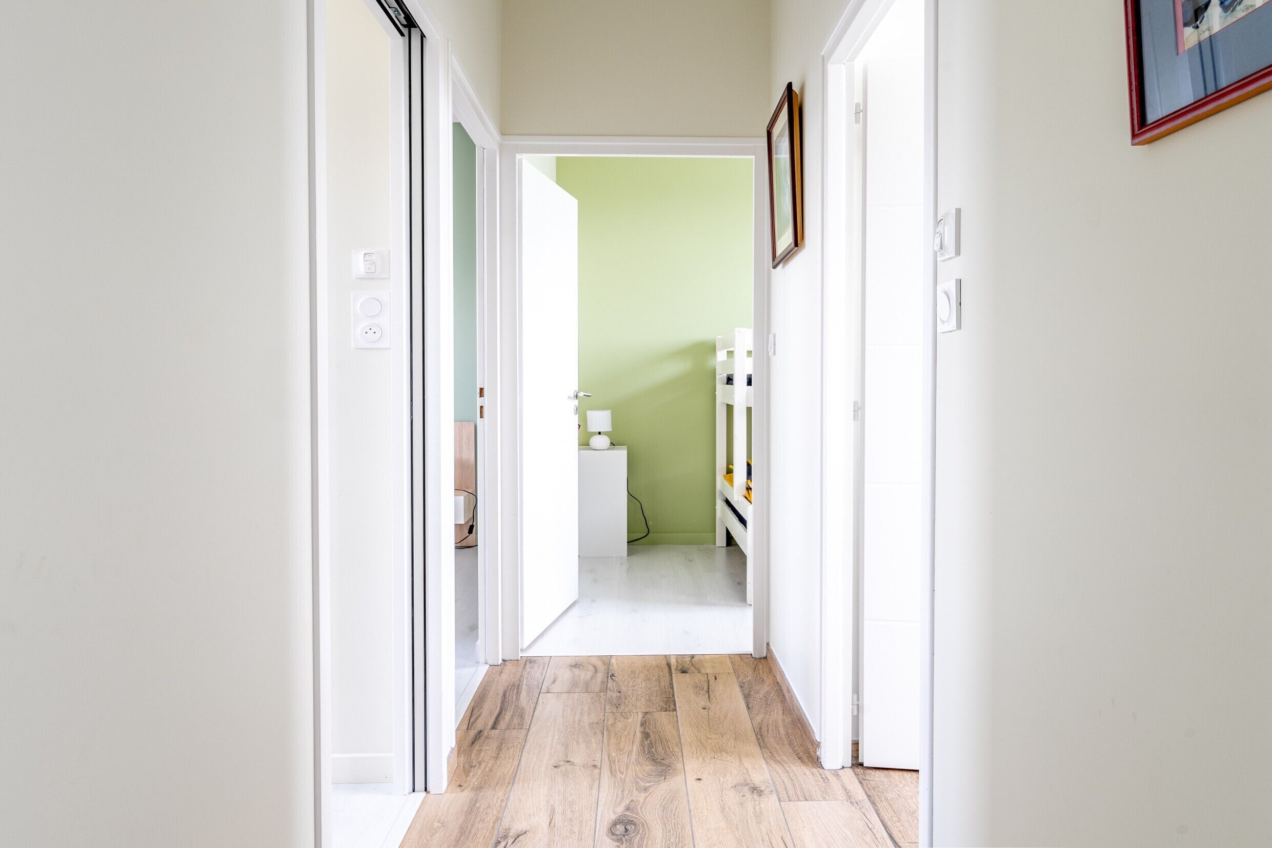 A hallway with white walls, a wooden floor, and a green room in the distance.