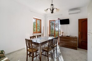 A dining area with a glass-top table, wooden chairs, a chandelier, a television, and a wooden cabinet.