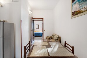 A narrow hallway leading to a bedroom with a bed, a dining table with chairs, a sofa, and a refrigerator.