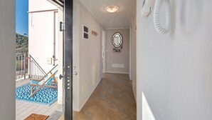 Un couloir doté d’un sol carrelé, d’un mur blanc orné d’un miroir et d’une porte donnant sur un espace extérieur agrémenté d’un paillasson bleu à motifs et d’une chaise.
