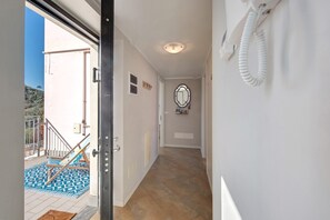 Um corredor com piso de azulejos, parede branca com espelho e porta que dá para área externa com tapete azul estampado e uma cadeira.