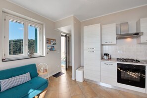 Eine moderne Küche mit Gasherd, Backofen und weißen Schränken. Ein Türkis Sofa mit gemustertem Kissen, ein kleiner Ventilator und ein Fenster mit Blick auf Grünflächen.