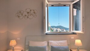Une chambre à coucher avec un lit, deux tables de chevet avec des lampes, une fenêtre donnant sur des bâtiments et des collines, et des œuvres d’art murales.
