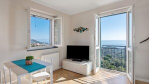 Une pièce dotée d’un téléviseur, d’une table avec des chaises, d’une plante en pot et offrant une vue sur la mer grâce à de grandes fenêtres.