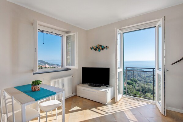 A room with a TV, a table with chairs, a potted plant, and a view of the sea through large windows.
