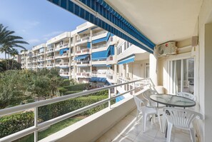 Un balcon doté d’une table en verre et de chaises blanches, donnant sur une cour verdoyante et un immeuble à plusieurs étages, lui aussi pourvu de balcons.