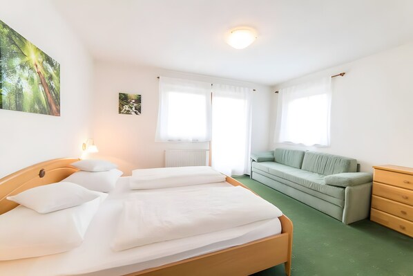 A bedroom with a wooden bed, a green sofa, a wooden dresser, and a window with white curtains.