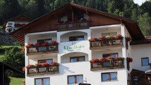 En bygning med flere etager, balkoner pyntet med blomster, et skilt der siger 'Klauenthaler Pension - Cafe,' og et bjergrigt landskab i baggrunden.