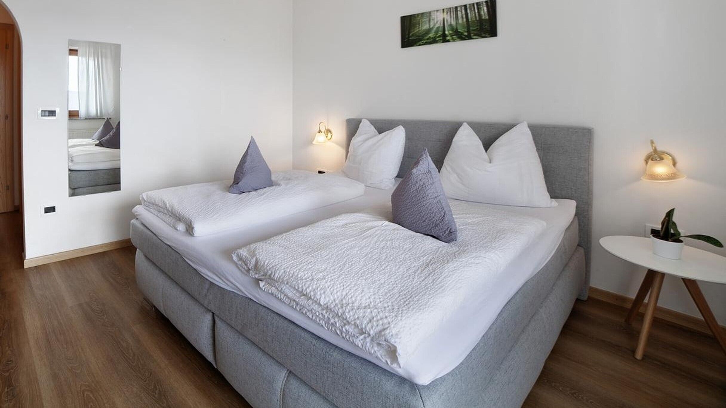 A double bed with a grey headboard, two bedside tables, a small round table with a plant, and a framed picture on the wall.