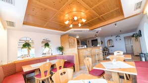 Een restaurant met houten tafels en rode, beklede zitplaatsen, een keuken en een plafond met zichtbare balken en een centrale lamp.