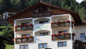Un immeuble à plusieurs étages, doté de balcons ornés de fleurs, une enseigne indiquant « Klauenthaler Pension - Café », et un paysage montagneux en arrière-plan.