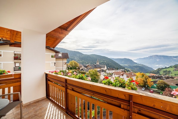 Un balcon doté de balustrades en bois, avec une table et des chaises, d’où l’on peut admirer un village, des maisons et des montagnes en arrière-plan.
