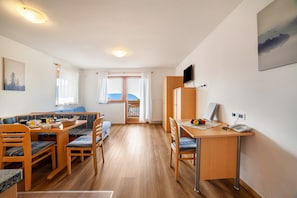 A room with a dining table, chairs, a desk with a telephone, and a television on the wall.