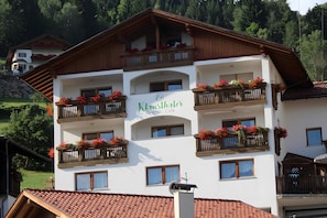 A multi-story building with balconies adorned with flowers, a sign reading 'Klauenthaler Pension - Cafe,' and a mountainous backdrop.
